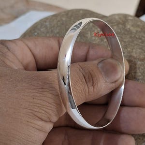 May include: A polished silver bangle bracelet. The smooth, rounded design reflects light, creating a bright, shiny appearance. The bracelet is a simple, elegant piece of jewelry.