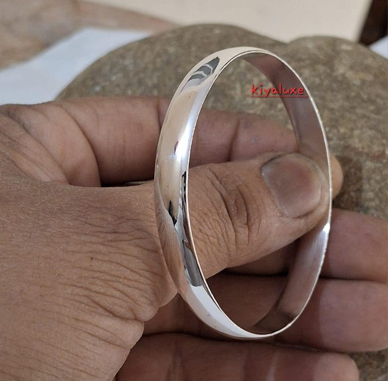 May include: A polished silver bangle bracelet. The smooth, rounded design reflects light, creating a bright, shiny appearance. The bracelet is a simple, elegant piece of jewelry.
