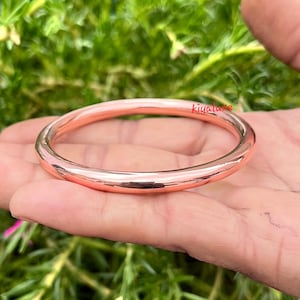 以下が含まれることがあります： 磨かれた銅製のバングルブレスレット。丸いブレスレットは、滑らかで反射性の表面を持つ、銅の無垢のバンドです。ブレスレットは、緑の葉の背景に対して手で保持されています。「kiyaluxe」という文字がブレスレットに表示されています。