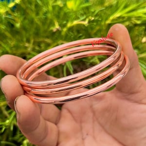 May include: Four copper-colored bangles are stacked together, reflecting light. The bangles are smooth and round, with a polished finish. The background is blurred green foliage, and the text "fiyaluxe" is visible.