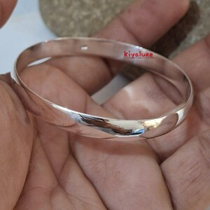 May include: A polished silver bangle bracelet with a smooth, rounded design. The bracelet reflects light, showcasing its shiny surface. The text "kivaluxe" is visible on the bracelet. This piece of jewelry is a classic accessory.