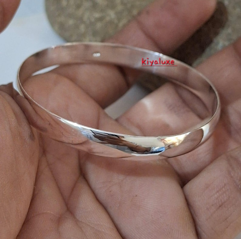 May include: A polished silver bangle bracelet with a smooth, rounded design. The bracelet reflects light, showcasing its shiny surface. The text "kivaluxe" is visible on the bracelet. This piece of jewelry is a classic accessory.