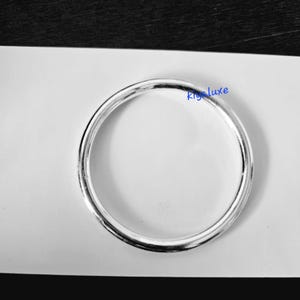 May include: A silver-colored, circular bracelet with a smooth, reflective surface. The bracelet is a simple, elegant design, suitable for everyday wear. The text "Kig luxe" is visible on the bracelet.