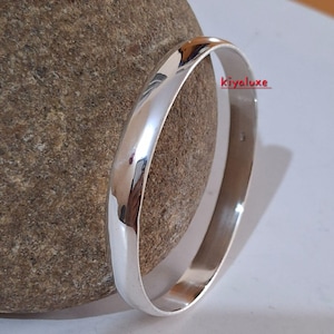 May include: A polished silver bangle bracelet with a smooth, rounded design. The bracelet reflects light, creating a bright, shiny appearance. The word "Kiyaluxe" is printed on the bracelet. This piece is a simple, elegant jewelry item.