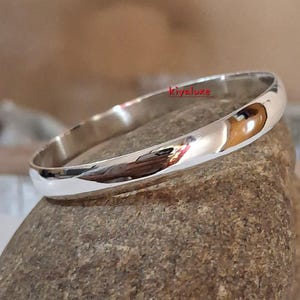 May include: A polished silver bangle bracelet. The smooth, rounded design reflects the surrounding environment. The bracelet is displayed on a textured, neutral-toned surface. The word "kivaluxe" is visible on the bracelet.