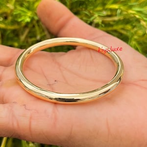 Könnte beinhalten: Ein goldfarbenes Armreif-Armband mit glattem, abgerundetem Design. Das Armband wird in einer Hand gehalten, und die Oberfläche reflektiert das Licht, wodurch seine polierte Oberfläche hervorgehoben wird. Das Wort "kivaluxe" ist auf dem Armband geschrieben.