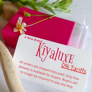 May include: A bright pink box with a white card featuring the brand name "Kiyaluxe" in red script. The card also states "A New Brand" and "0% Tariffs". A small white flower and green leaves are on top of the box.