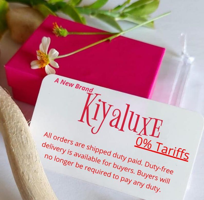 May include: A bright pink box with a white card featuring the brand name "Kiyaluxe" in red script. The card also states "A New Brand" and "0% Tariffs". A small white flower and green leaves are on top of the box.