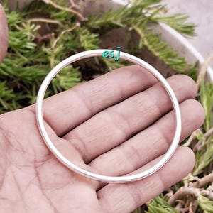 May include: A silver-colored bangle bracelet. The circular bracelet is held in a hand, with green foliage in the background. The text "esj" is visible in the upper right corner.