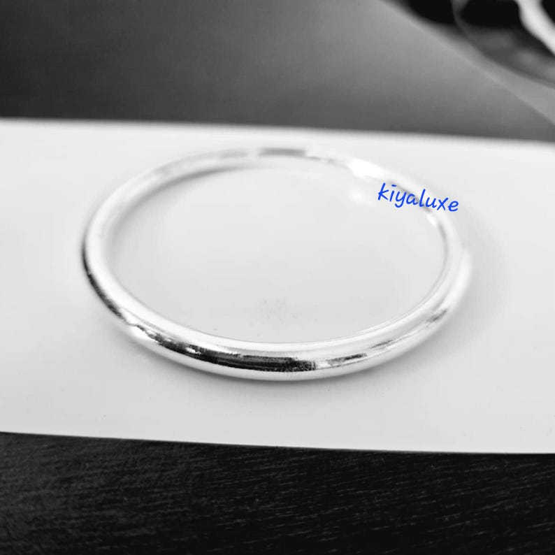 May include: A silver ring with a smooth, rounded design. The ring is circular and sits on a white surface. The text "kiyaluxe" is visible on the ring. This ring is a piece of jewelry.