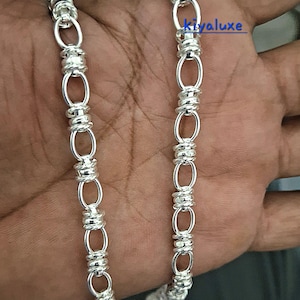 May include: A silver chain necklace featuring a repeating pattern of elongated oval links alternating with segments of tightly coiled silver wire. The text kiyaluxe is visible in blue near the top right of the metallic jewelry piece.