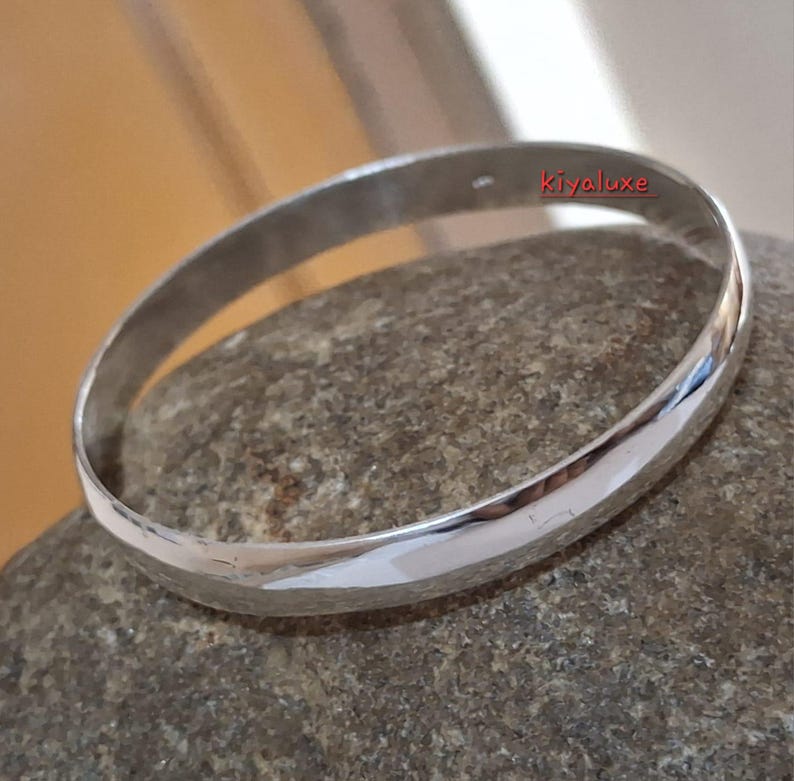May include: A polished silver bangle bracelet. The smooth, rounded bracelet reflects light, creating a bright, shiny appearance. The bracelet is resting on a gray stone. The text "Kiyaluxe" is visible on the bracelet.