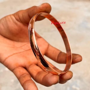 May include: A polished copper bangle bracelet. The smooth, rounded bracelet reflects light, showcasing its warm, reddish-brown color. The bracelet is held in a hand, with the text "Kiyaluxe" in pink.