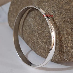 May include: A shiny, silver-colored bangle bracelet. The smooth, rounded band reflects light, creating highlights and shadows. The bracelet is displayed on a textured, gray-brown stone, showcasing its polished surface. A simple, elegant piece of jewelry.