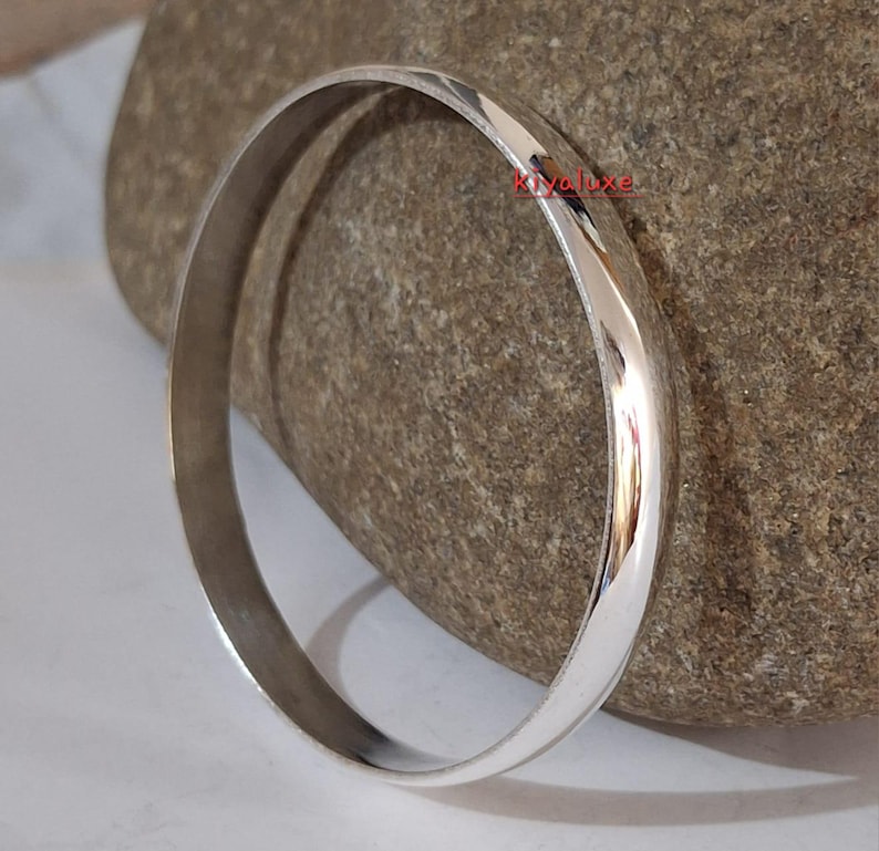 May include: A shiny, silver-colored bangle bracelet. The smooth, rounded band reflects light, creating highlights and shadows. The bracelet is displayed on a textured, gray-brown stone, showcasing its polished surface. A simple, elegant piece of jewelry.