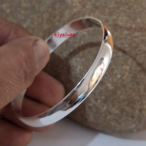 May include: A polished silver bangle bracelet. The smooth, rounded design reflects light, creating a bright, shiny appearance. The bracelet is held in a hand, with the text "kiyaluxe" visible on the bracelet.