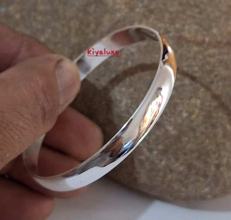 May include: A polished silver bangle bracelet. The smooth, rounded design reflects light, creating a bright, shiny appearance. The bracelet is held in a hand, with the text "kiyaluxe" visible on the bracelet.