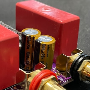 Op de afbeelding: Close-up van elektronische componenten op een printplaat. De afbeelding toont twee cilindrische condensatoren met goudzwarte labels, een rood rechthoekig component en goudkleurige RCA-connectoren met rode en zwarte ringen.