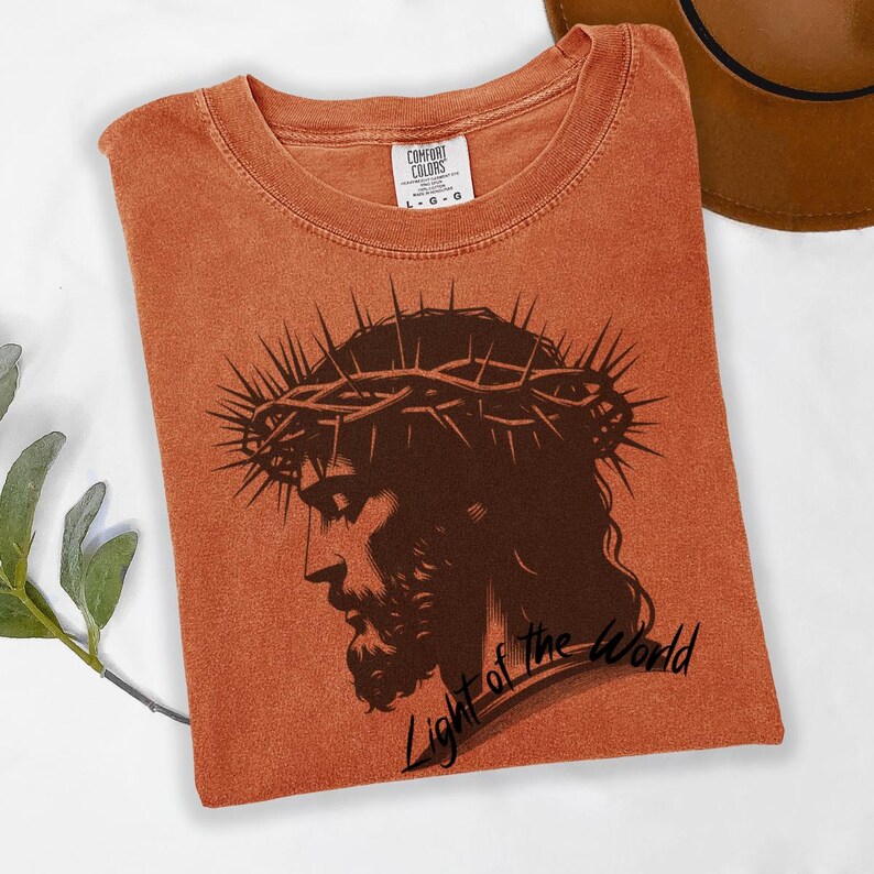 May include: A burnt orange t-shirt featuring a black and white image of Jesus wearing a crown of thorns. The text "Light of the World" is printed below the image.