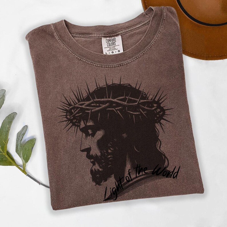 May include: A brown t-shirt featuring a black and white image of Jesus wearing a crown of thorns. The text "Light of the World" is printed below the image.