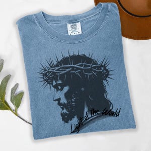 May include: A light blue t-shirt featuring a black and white image of Jesus wearing a crown of thorns. The text "Light of the World" is written below the image.