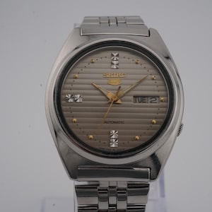 Puede incluir: Reloj de pulsera automático Seiko plateado con esfera gris y correa de acero inoxidable a juego. La esfera del reloj presenta agujas e indicadores dorados, con el día mostrado a las 3 en punto. El reloj se muestra sobre un soporte transparente.