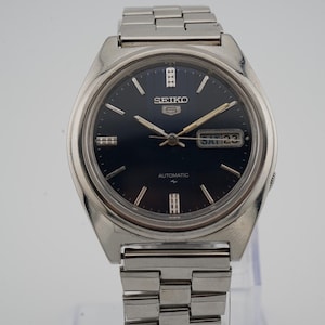 Puede incluir: Un reloj de pulsera automático Seiko 5 plateado con esfera azul oscuro. El reloj presenta una pantalla de día y fecha, agujas plateadas y una correa de eslabones plateada. El reloj se exhibe sobre un soporte transparente.