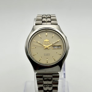 Puede incluir: Reloj de pulsera Orient plateado con esfera color crema. El reloj presenta agujas e indicadores horarios dorados, y una pantalla de día y fecha. La correa del reloj es de metal. El reloj se exhibe sobre un soporte transparente.