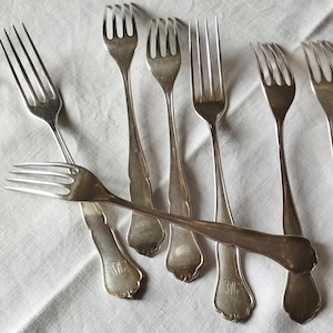 Peut inclure: Ensemble de fourchettes en argent ancien disposées sur une nappe en lin blanc. Les fourchettes présentent un design classique à trois dents et des manches ornés. La nappe comporte un monogramme brodé dans le coin.