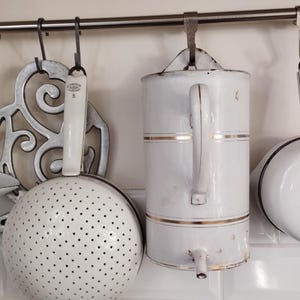 Può includere: Utensili da cucina in smalto bianco appesi a un rack in metallo. Gli articoli includono un colino, una caraffa e una pentola. Il rack è montato su una parete piastrellata bianca.