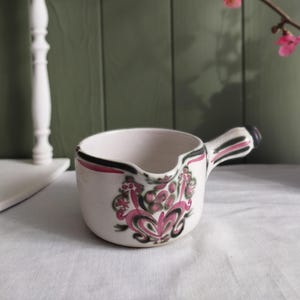 May include: A small, white ceramic creamer with a handle and a spout. The creamer is decorated with a pink and green floral design and black and pink trim. A sprig of pink flowers is in the background.