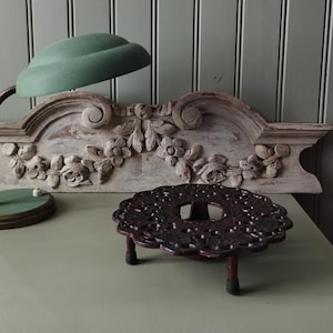 Op de afbeelding: Een groene bureaulamp met een gebogen kap en een decoratief, gesneden, wit houten hoofdbord met bloemendetails. Een donkerrode, gietijzeren onderzetter met een bloemmotief.