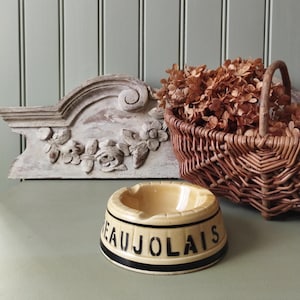 Op de afbeelding: Een gele asbak van keramiek met zwarte letters die "BEAUJOLAIS" lezen.