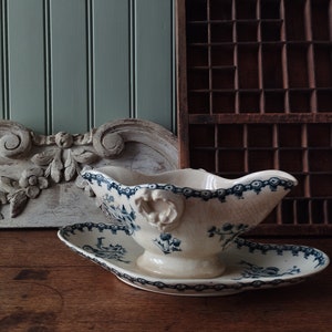 May include: A cream-coloured ceramic gravy boat with a matching oval underplate, featuring a blue floral transferware pattern, a scalloped rim, and decorative moulded handles on the sides, showcasing a visible crackle glaze finish throughout.
