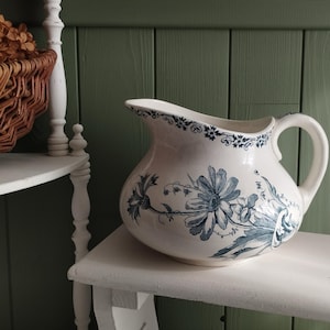 Può includere: Una brocca in ceramica color crema con un motivo floreale blu a trasferimento. Il design presenta grandi fiori simili a margherite e fogliame sul corpo, con un bordo floreale decorativo lungo l'apertura e un manico curvo sul lato destro.