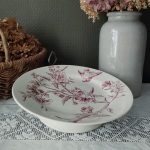 Peut inclure: Plat de service en céramique blanche avec un bord surélevé, orné d'un motif floral et d'oiseaux bordeaux. Le plat est posé sur une nappe en dentelle blanche, avec un panier de fleurs séchées et un vase blanc en arrière-plan.