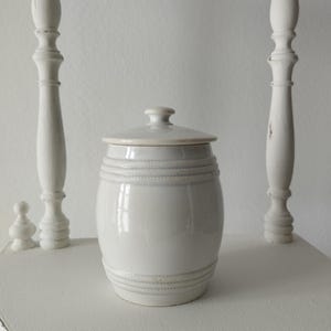 Op de afbeelding: Een witte, tonvormige keramische pot met een bijpassend deksel. De pot heeft een gladde, glanzende afwerking en is versierd met twee horizontale banden met een verhoogde touwstructuur die de boven- en onderkant van het lichaam omcirkelen.