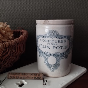 以下が含まれることがあります： 青い文字で「CONFITURES FINES FÉLIX POTIN」と書かれたヴィンテージの陶器の瓶。瓶はオフホワイトで蓋付きで、白い正方形のタイルに置かれています。テキストとワイヤーの付いたコルクが瓶の前にあります。背景には籐のバスケットがあります。