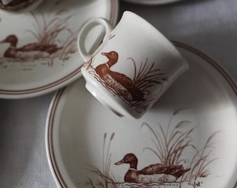 Op de afbeelding: Een set off-white keramieken servies met een bruin transferpatroon van eenden die in een moeras met hoog riet zwemmen. De collectie bevat meerdere borden en kopjes, elk versierd met dezelfde gedetailleerde illustratie van in het wild levende dieren.