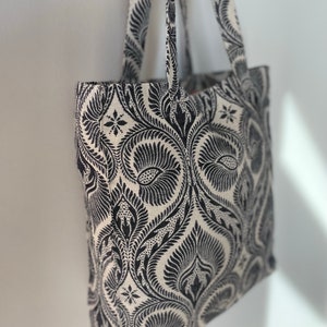 K&ouml;nnte beinhalten: Eine schwarze und wei&szlig;e Einkaufstasche mit einem Blumenmuster. Die Tasche hat zwei Henkel und ist aus Stoff.