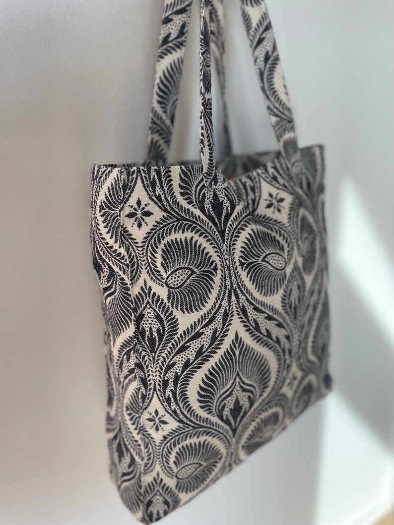 K&ouml;nnte beinhalten: Eine schwarze und wei&szlig;e Einkaufstasche mit einem Blumenmuster. Die Tasche hat zwei Henkel und ist aus Stoff.