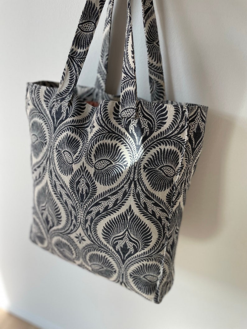 K&ouml;nnte beinhalten: Eine schwarze und wei&szlig;e Einkaufstasche mit einem Blumenmuster. Die Tasche hat zwei Henkel und ist aus robustem Stoff.