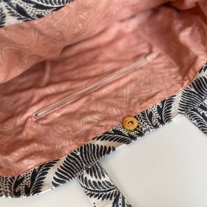 K&ouml;nnte beinhalten: Eine schwarz-wei&szlig;e Einkaufstasche mit floralem Muster. Die Tasche hat eine Rei&szlig;verschlusstasche und einen Druckknopfverschluss. Das Innere der Tasche ist hellrosa.