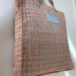 K&ouml;nnte beinhalten: Eine hellbraune Tragetasche mit einem wei&szlig;en und braunen geometrischen Muster. Die Tasche hat einen blauen Aufn&auml;her mit dem Text "Willkommen in meinem Abenteuer" darauf.