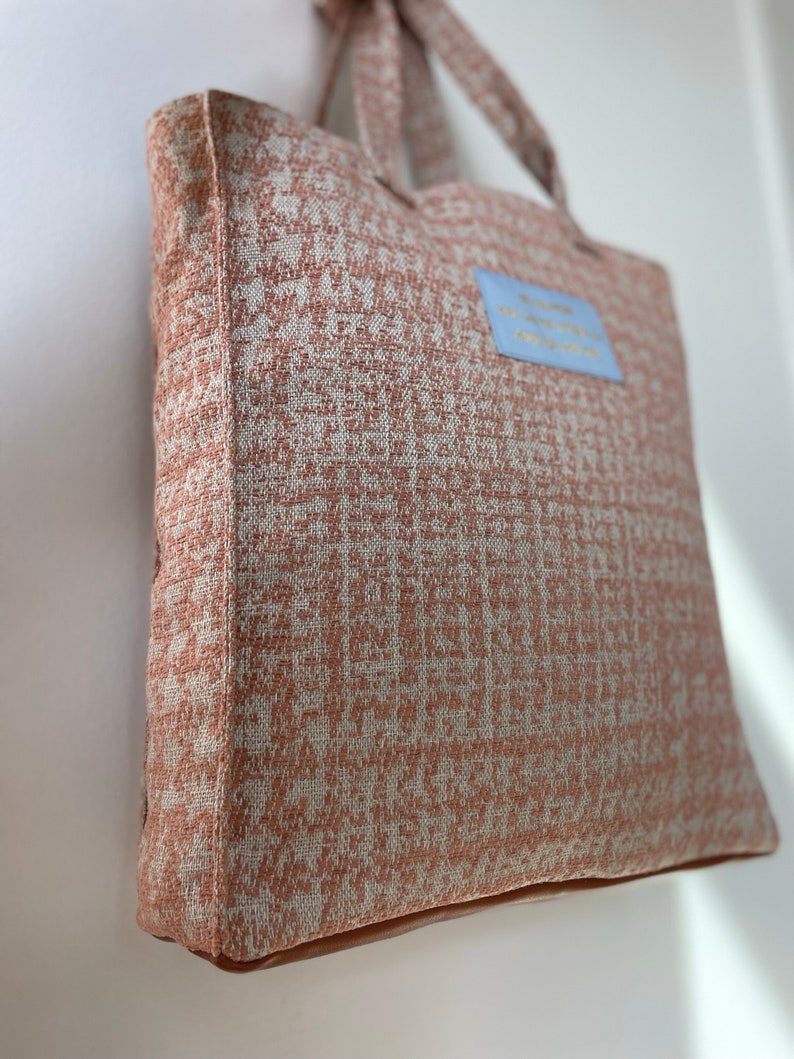 K&ouml;nnte beinhalten: Eine hellbraune Tragetasche mit einem wei&szlig;en und braunen geometrischen Muster. Die Tasche hat einen blauen Aufn&auml;her mit dem Text "Willkommen in meinem Abenteuer" darauf.