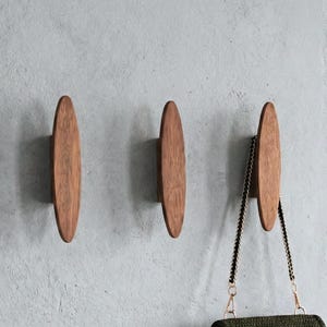 May include: Three oval wooden wall hooks are fixed to a grey wall. A dark green crocheted handbag with a gold clasp hangs from one of the hooks. A small succulent plant in a grey pot sits on a wooden shelf.