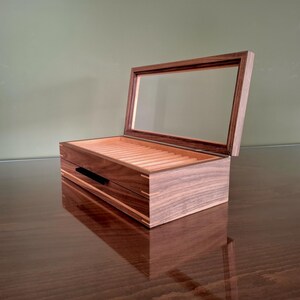 May include: A rectangular wooden display box featuring a dark brown walnut exterior with light wood corner joinery. The box has a hinged lid with a clear glass panel and an interior lined with light-colored wood featuring multiple parallel grooves.