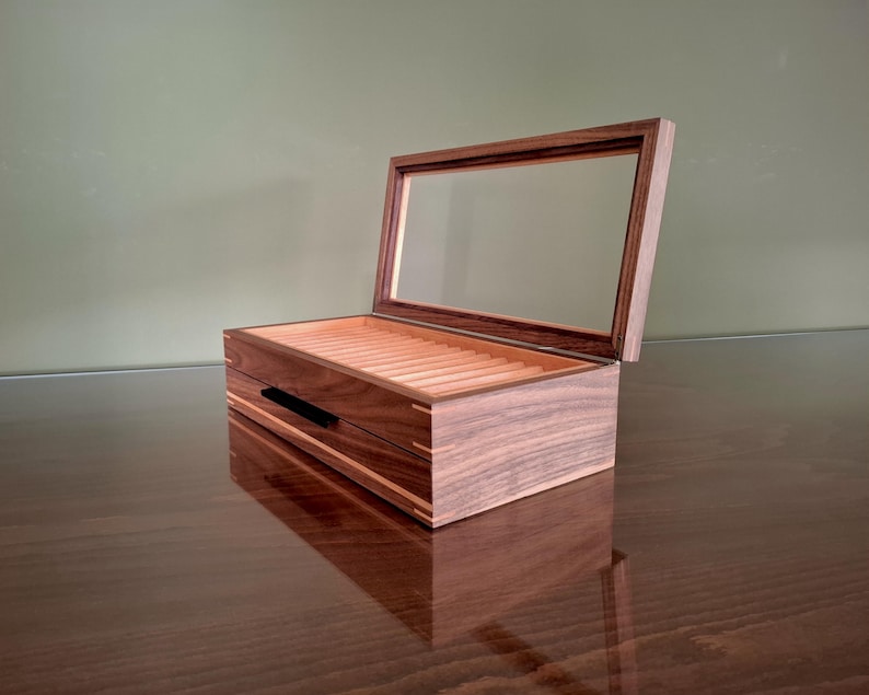 May include: A rectangular wooden display box featuring a dark brown walnut exterior with light wood corner joinery. The box has a hinged lid with a clear glass panel and an interior lined with light-colored wood featuring multiple parallel grooves.