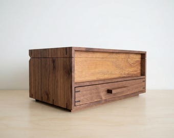 May include: A rectangular wooden storage box featuring a dark walnut frame with visible joinery details. The front includes a single horizontal drawer with a thin, protruding wooden handle and a lighter-toned wood panel section above the drawer.