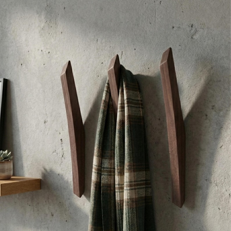 Dark walnut mid-century modern wooden wall hooks mounted on a textured light gray wall, with a cozy plaid scarf hanging from the center hook, styled in a minimalist home interior with a small wooden shelf and potted plant